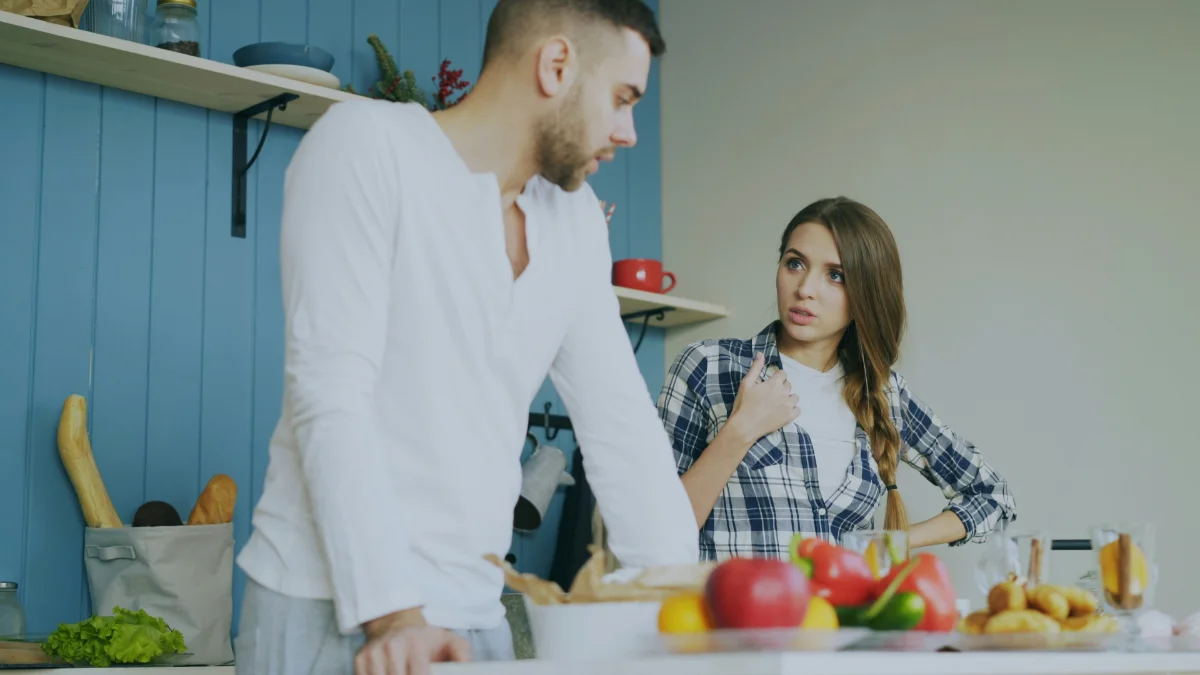A couple in their kitchen mid-disagreement, he turned away from her, she watching him with a hand raised