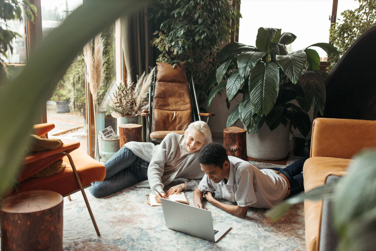 Two people lying on the floor of a sunlit, plant-filled room, working through the course together with a laptop and an open notebook
